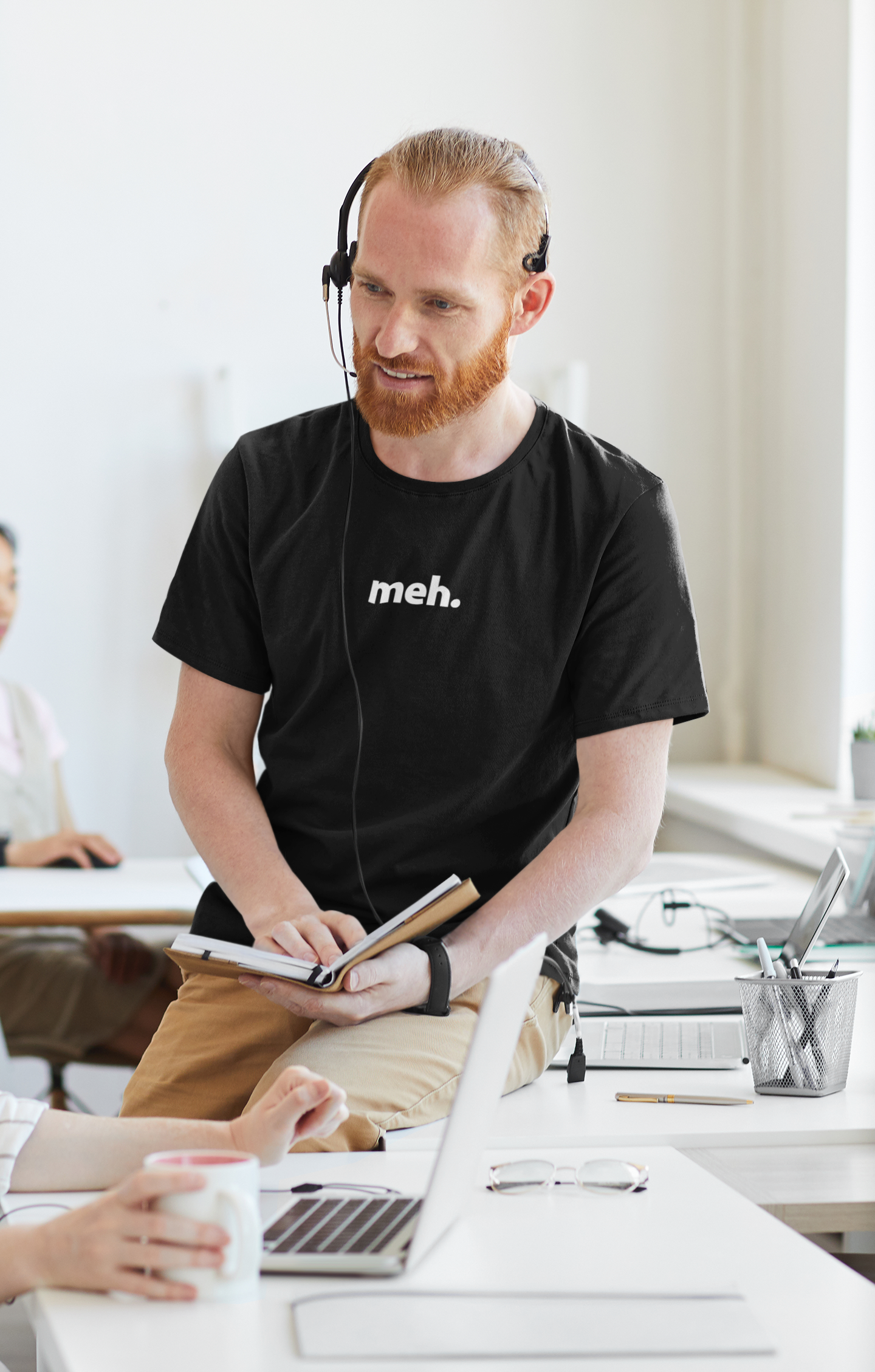 Man wearing a black t-shirt with 'meh.' text, sitting at a desk in an office setting.