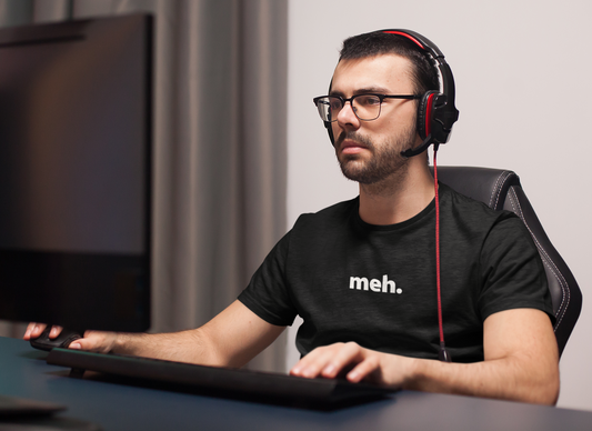 Man wearing headphones and a black t-shirt with 'meh.' text, sitting at a desk.